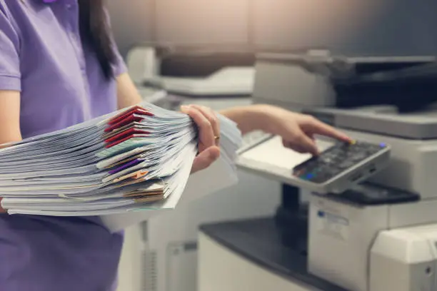 Photo of Bussinesswoman using copier machine to copy heap of paperwork in office.