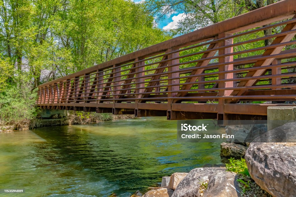 Bridge over glistening river with rocks on the bank at Ogden River Parkway Bridge over glistening river with rocks on the bank at Ogden River Parkway. The bridge has an amazing view of abundant trees and blue sky with puffy clouds. Ogden - Utah Stock Photo