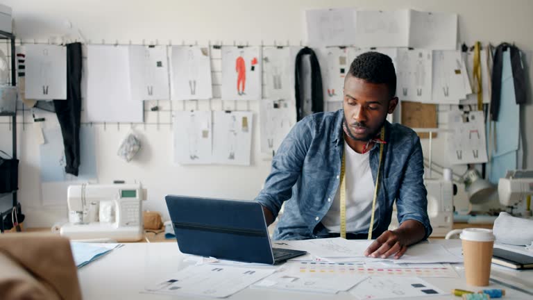 Bearded man designer drawing clothes sketching using laptop in modern studio