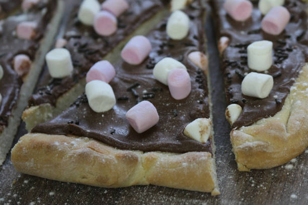 Stock photo of marshmallow and chocolate pizza cake, unhealthy diet eating mini marshmallows and hot chocolate pizza served for family / kid's birthday party, high in calories and taste imagen de pastel de pizza de chocolate casero con ingredientes de malvaviscos, rociados y azúcar glas, corteza de pizza horneada llena de avellanas y rematada con caramelos de chocolate caliente de mini malvavisco rojo y blanco, listo para el pastel de fi