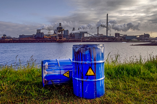 Empty Blue Chemical Waste Drums Lying on an Abandoned Bank with a view on Smoking Exhaust Pipes of a Heavy Industrial Factory