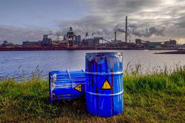 chemical waste drums in front of heavy industry Empty Blue Chemical Waste Drums Lying on an Abandoned Bank with a view on Smoking Exhaust Pipes of a Heavy Industrial Factory drum container stock pictures, royalty-free photos & images