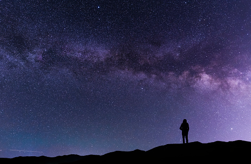 A silhouette stands on in the hill and looks at the Milky Way galaxy.