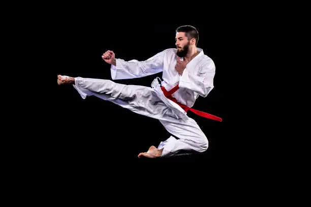 Male karate practitioner in mid-air during a flying kick. Black background.