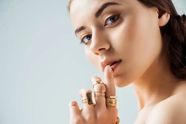 young woman with shiny makeup and golden rings touching lips isolated on grey