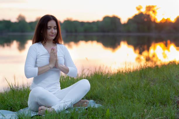 young woman in white clothes doing yoga, hands at the heart chakra in front of the chest namaste by the lake at sunset, sitting on a lush meadow in the lotus position stock photo
