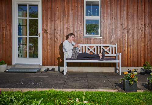 Woman knitting resting on bench. Her home is a detached house in the countryside with garden. House is made of wood and painted brown.