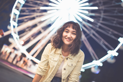 Beautiful asian girl in an amusement park at nght, smiling. Ferris wheel in the background. Copy space.