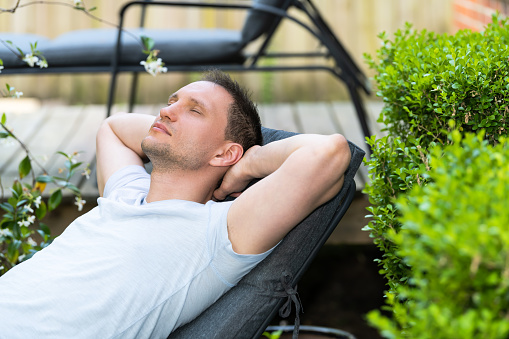 Young man sitting leaning back on patio lounge chair sleeping in outdoor spring flower garden in backyard of home plants