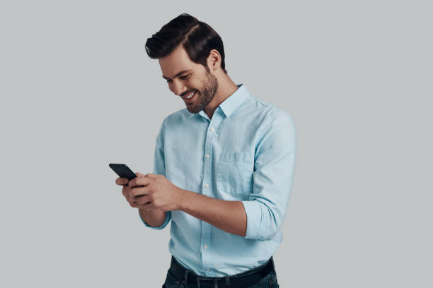 istock Chat with friend. Handsome young man using smart phone and smiling while standing against grey background 1153664533