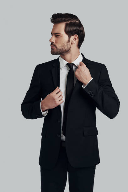 istock Confidence and charisma. Handsome young man in full suit adjusting jacket and looking away while standing against grey background 1153664670