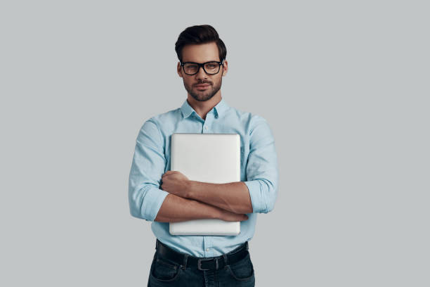istock Confident and smart. Handsome young man carrying laptop and smiling while standing against grey background 1153664708