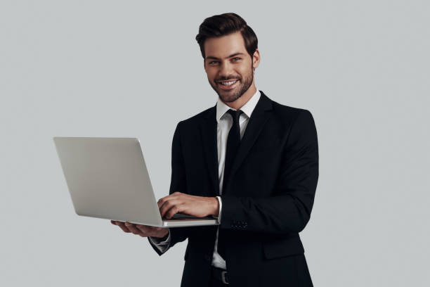 istock Confident business expert. Handsome young man in full suit looking at camera and using laptop while standing against grey background 1153665316