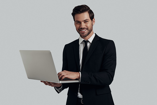 Confident business expert. Handsome young man in full suit looking at camera and using laptop while standing against grey background