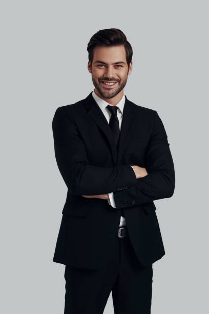 istock Confident business expert. Handsome young man in full suit smiling and looking at camera while standing against grey background 1153665385
