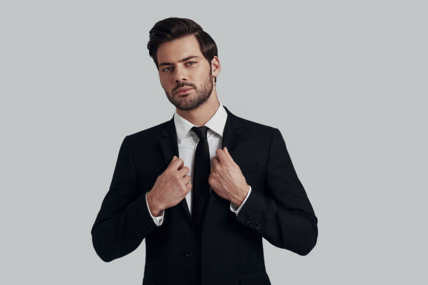 istock Confident manager. Handsome young man in full suit adjusting jacket and looking at camera while standing against grey background 1153665518