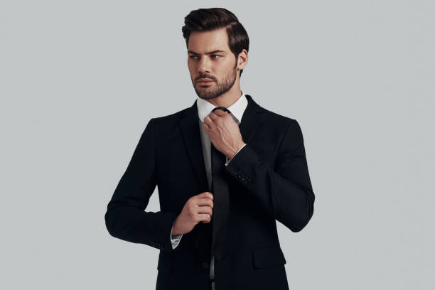 istock Everything must be perfect. Handsome young man in full suit adjusting tie and looking away while standing against grey background 1153665593