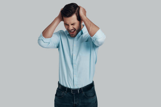 istock Failure. Frustrated young man keeping head in hands and making a face while standing against grey background 1153665628