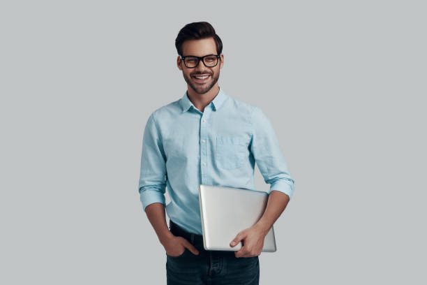 istock Happy businessman. Handsome young man carrying laptop and looking at camera while standing against grey background 1153666097