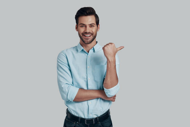 istock Look over there! Handsome young man looking at camera with smile and pointing copy space while standing against grey background 1153667280