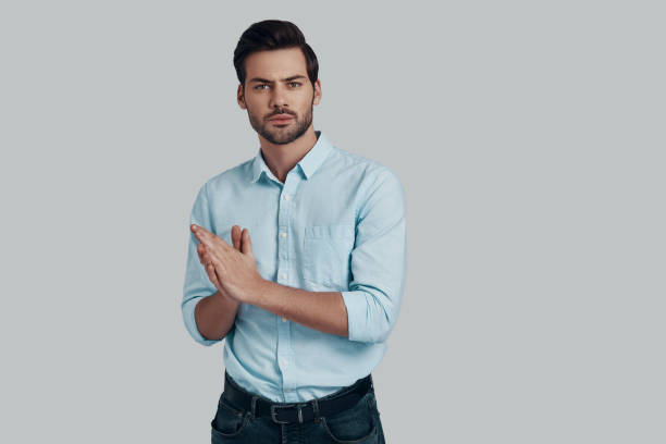 istock Lost in thoughts. Handsome young man looking at camera and keeping hands clasped while standing against grey background 1153667385