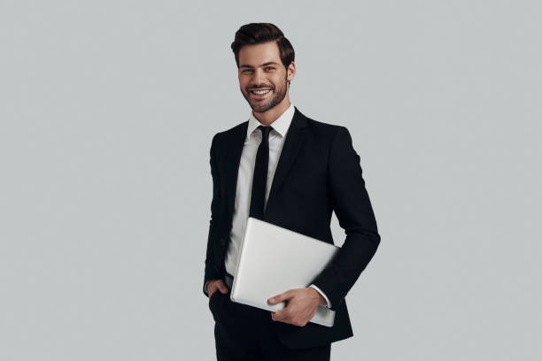 istock Modern businessman. Handsome young man in full suit carrying laptop while standing against grey background 1153667509