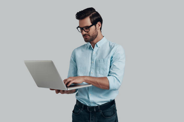 istock Paying attention to every detail. Young man using laptop while standing against grey background 1153667645