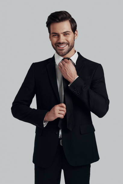 istock Perfect style. Handsome young man in full suit adjusting tie and looking at camera with smile while standing against grey background 1153667971