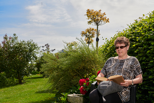 Retired people minding their garden. Having gardening as hobby after retirement. Fresh and green garden with hedge and flowerbeds. Woman reading