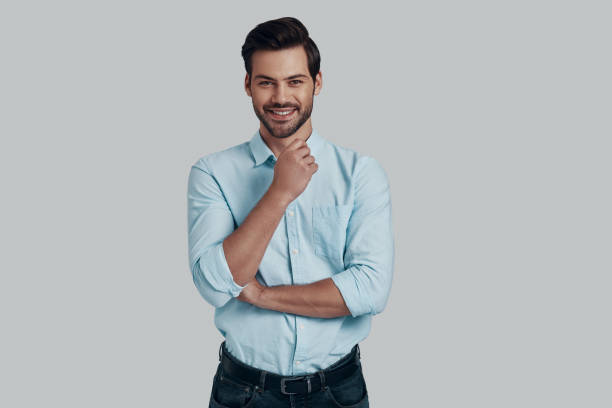istock Pure confidence. Handsome young man looking at camera and keeping hand on chin while standing against grey background 1153668124