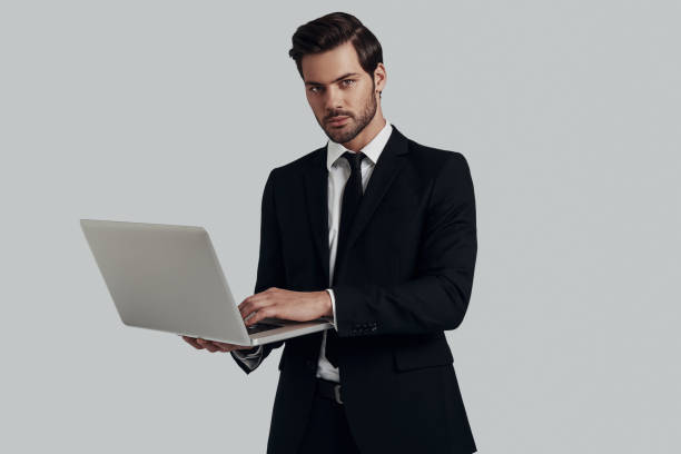 istock Ready to help. Handsome young man in full suit looking at camera and using laptop while standing against grey background 1153668245