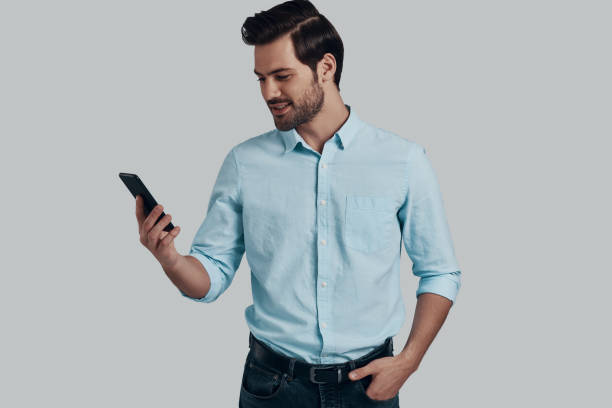 istock Receiving messages from friends. Handsome young man using smart phone and smiling while standing against grey background 1153668296