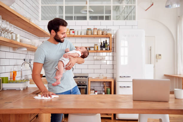 Multi-Tasking Father Holds Sleeping Baby Son And Cleans In Kitchen Multi-Tasking Father Holds Sleeping Baby Son And Cleans In Kitchen father housework stock pictures, royalty-free photos & images