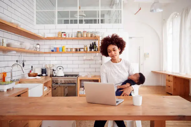 Photo of Multi-Tasking Mother Holds Sleeping Baby Son And Works On Laptop Computer In Kitchen