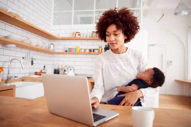 Photo of Multi-Tasking Mother Holds Sleeping Baby Son And Works On Laptop Computer In Kitchen