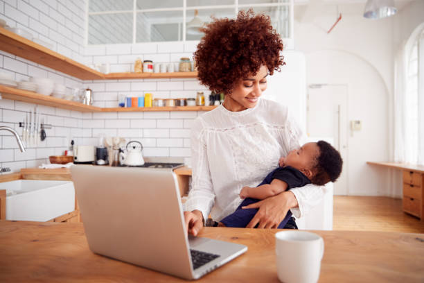 istock Multi-Tasking Mother Holds Sleeping Baby Son And Works On Laptop Computer In Kitchen 1153669114