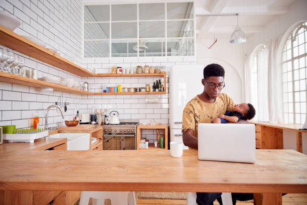 istock Multi-Tasking Father Holds Sleeping Baby Son And Works On Laptop Computer In Kitchen 1153669379