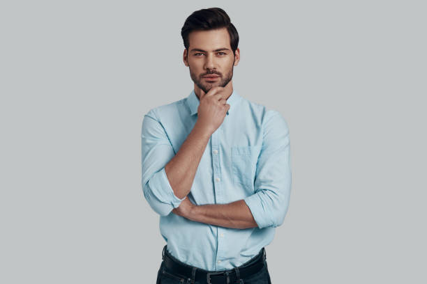 istock What if... Handsome young man looking at camera and keeping hand on chin while standing against grey background 1153669997