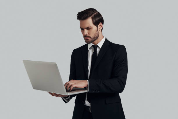 istock Working on the go. Handsome young man in full suit using laptop while standing against grey background 1153670041