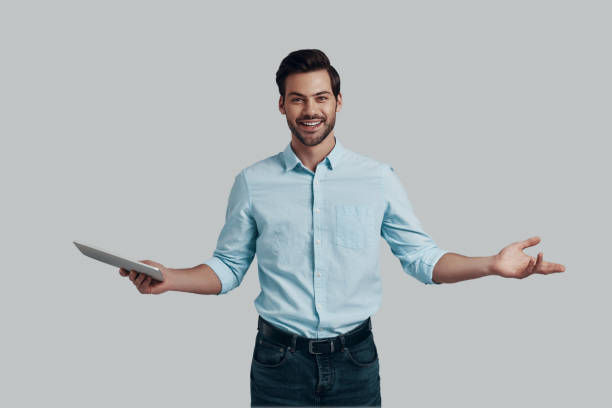 istock You are welcome! Handsome young man keeping arms outstretched and looking at camera while standing against grey background 1153670706