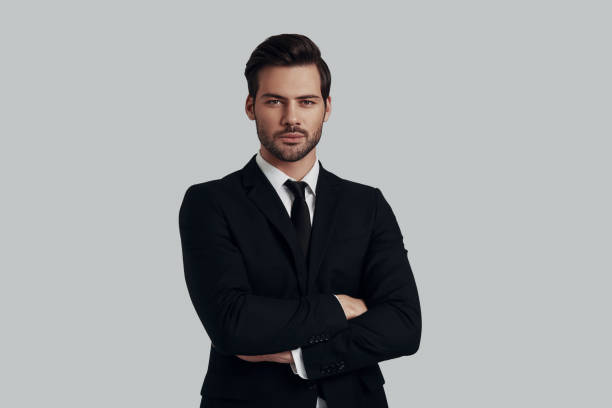 istock Young and confident. Handsome young man in full suit looking at camera and keeping arms crossed while standing against grey background 1153670844