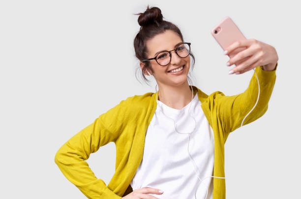 Pretty young European woman in casual clothes standing and taking a selfie isolated over white studio background. Pretty Caucasian brunette female wearing transparent spectacles making self portrait stock photo