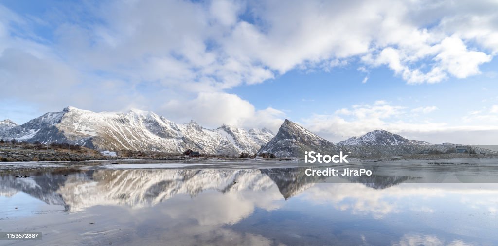 Panorama reflection of snowy mountains, blue sky and clouds on the calm clear water on a beach. Panorama reflection of snowy mountains, blue sky and clouds on the calm clear water on a beach. A range of pointy mountain in beautiful winter landscape of Lofoten in northern Norway. Desolated land. Purity Stock Photo