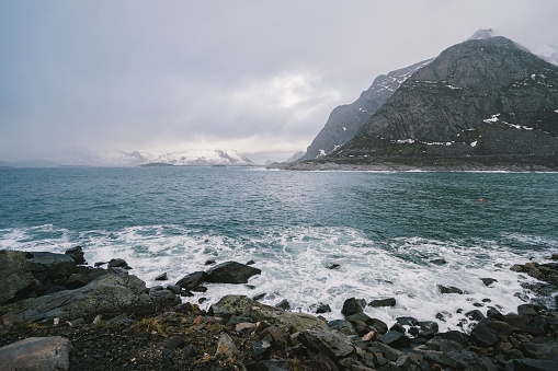 Waves are breaking on the shore and foaming dark blue turquoise sea water on the shoreline under cloudy rainy sky. Big mountain on the background.