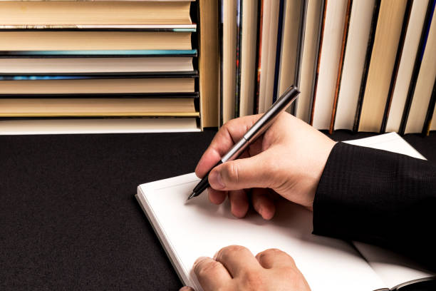 Hand holding pen over white pad of paper with blue lines with black background