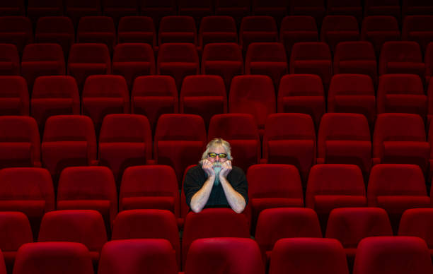 One man with white beard sits in Empty cinema or theatre with comfortable red seats stock photo