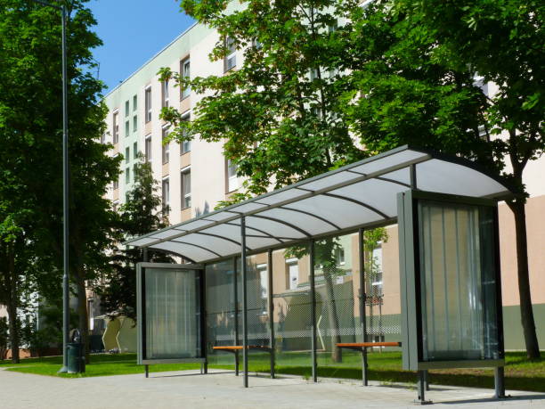 modern bus shelter design. multi unit apartment in the background bus shelter at a bus stop of glass and aluminum structure in park-like setting in day time with green background and appealing polka dot safety glass design and wooden benches & poster display glass bus hungary stock pictures, royalty-free photos & images
