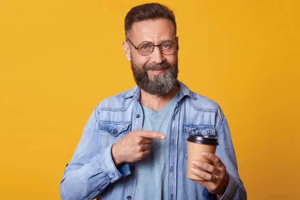 Delighted positive smiling handsome guy holding papercup of strong coffee in one hand, pointing at it with forefinger, having lunch, enjoying rest , wearing fashionable accessorie, looks satisfied.