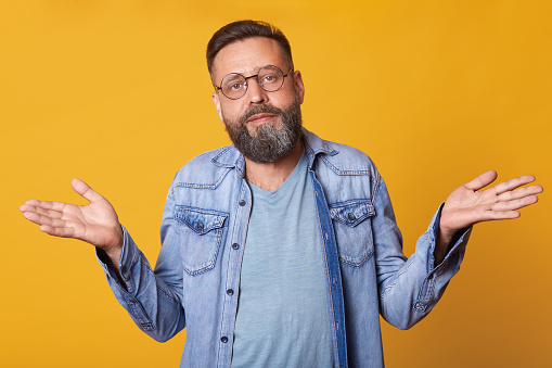 Hopeless desperate middle aged man wearing blue casual t shirt, jeans jacket and fashionable round spectacles, throwing hands up, being confused, not knowing what to do. People and emotions concept.