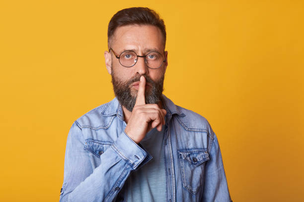 istock Serious middle aged emotionless bearded man in blue shirt, showing silence gesture, asks to keep secret, putting forefinger on lips, male dressed in denim jacket stands over pink background. 1153686308